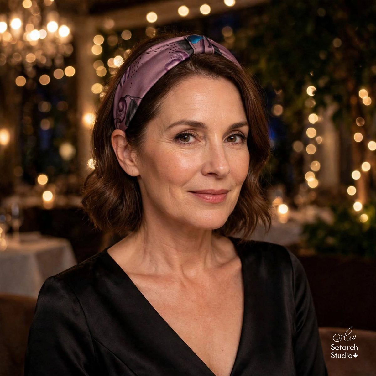 Real photo: a woman wearing the Seeds in the Twilight headband, botanical seed illustrations on dusty mauve, at an evening restaurant with warm string lights