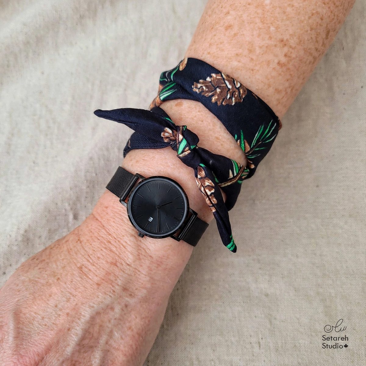 Real photo: the Nocturnal Pine Forest twilly worn as a bracelet beside a black watch, pine cones and green pine needles on dark navy