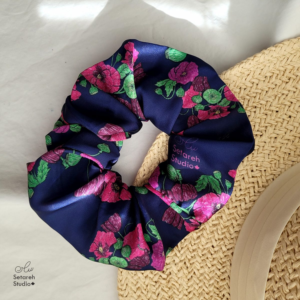 Real photo: the Midnight Poppy Garden scrunchie in a flat-lay beside a woven straw hat, vivid pink poppies on dark navy